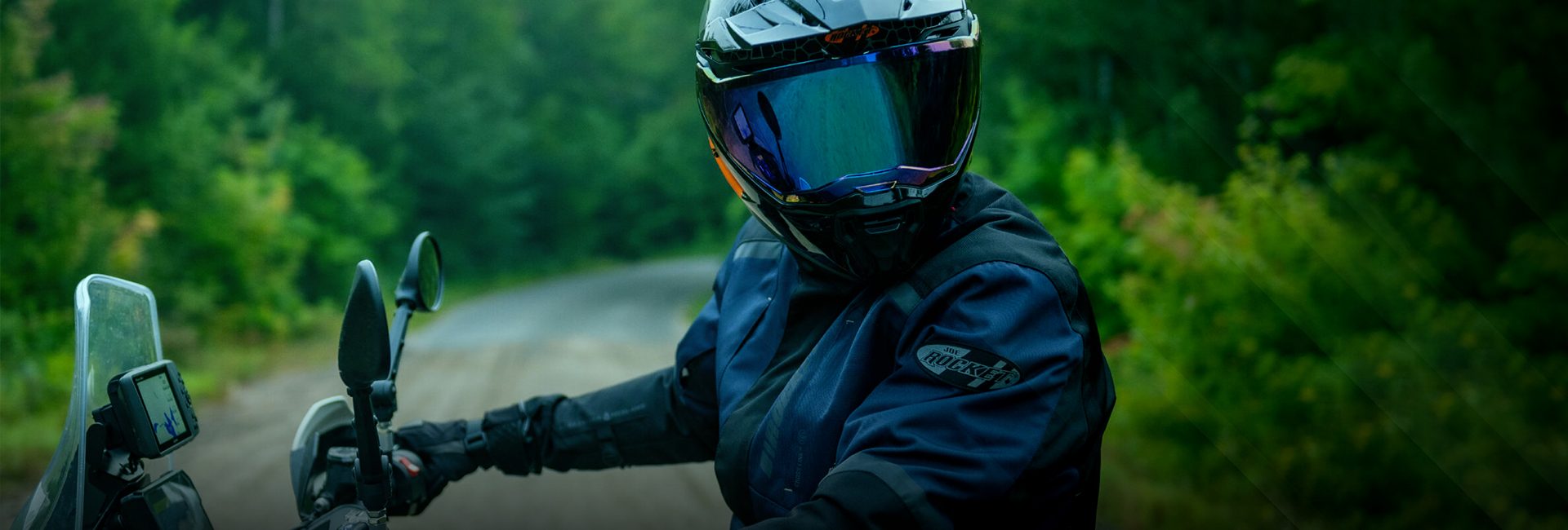 Motociclista con casco integral y chamarra Joe Rocket conduciendo por camino de terracería rodeado de naturaleza.