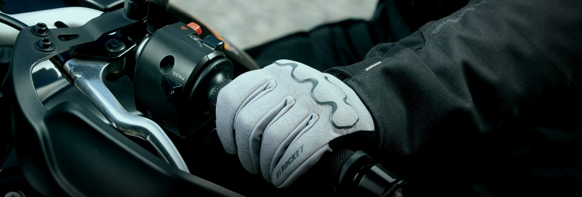Primer plano de una motociclista usando guantes blancos Joe Rocket mientras sujeta el manillar y la palanca de freno de una motocicleta.