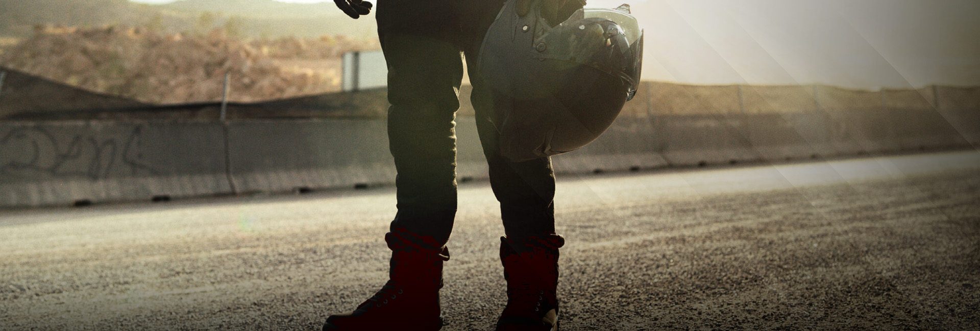 Motociclista en carretera al atardecer, con botas negras y pantalón de protección, sosteniendo casco.